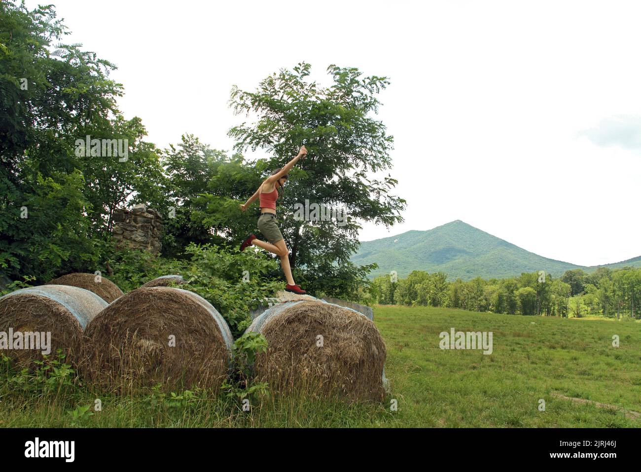 Summer landscape with hay bales in a field at the foot of the Blue Ridge Mountains, VA, USA. Girl jumping over hay bales. Stock Photo