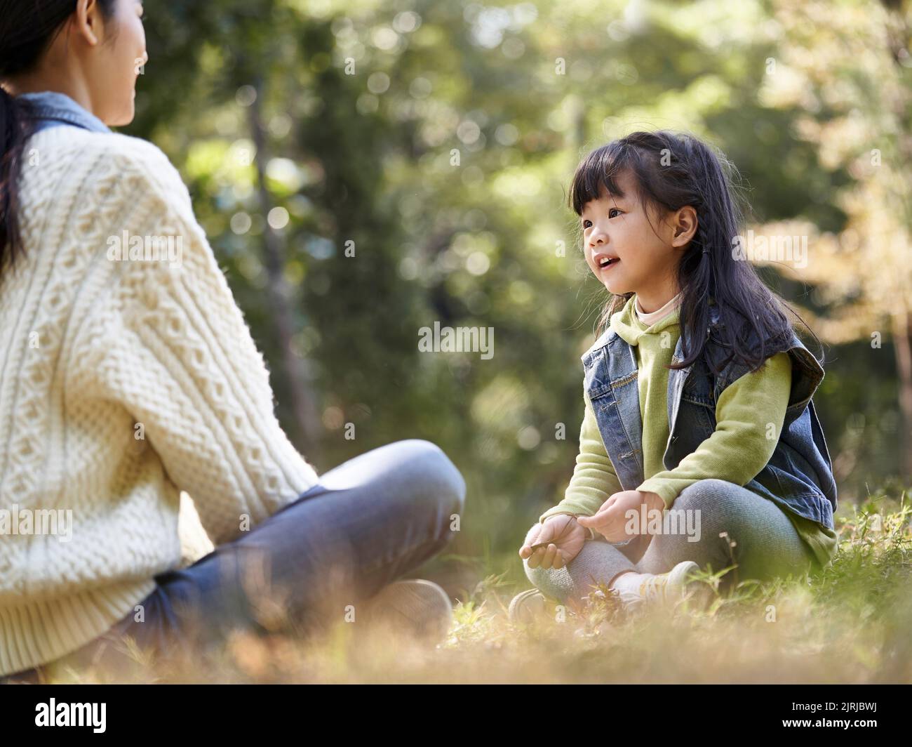 young asian mother sitting on grass in park having a pleasant conversation with preschool daughter Stock Photo