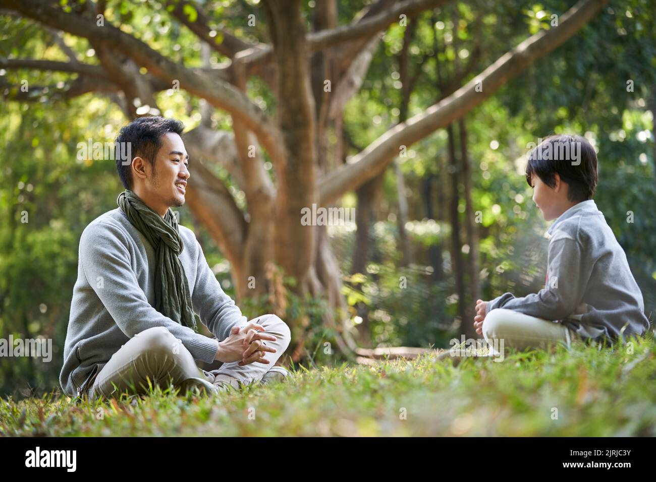 asian father and son sitting on grass having a pleasant conversation outdoors in park Stock Photo