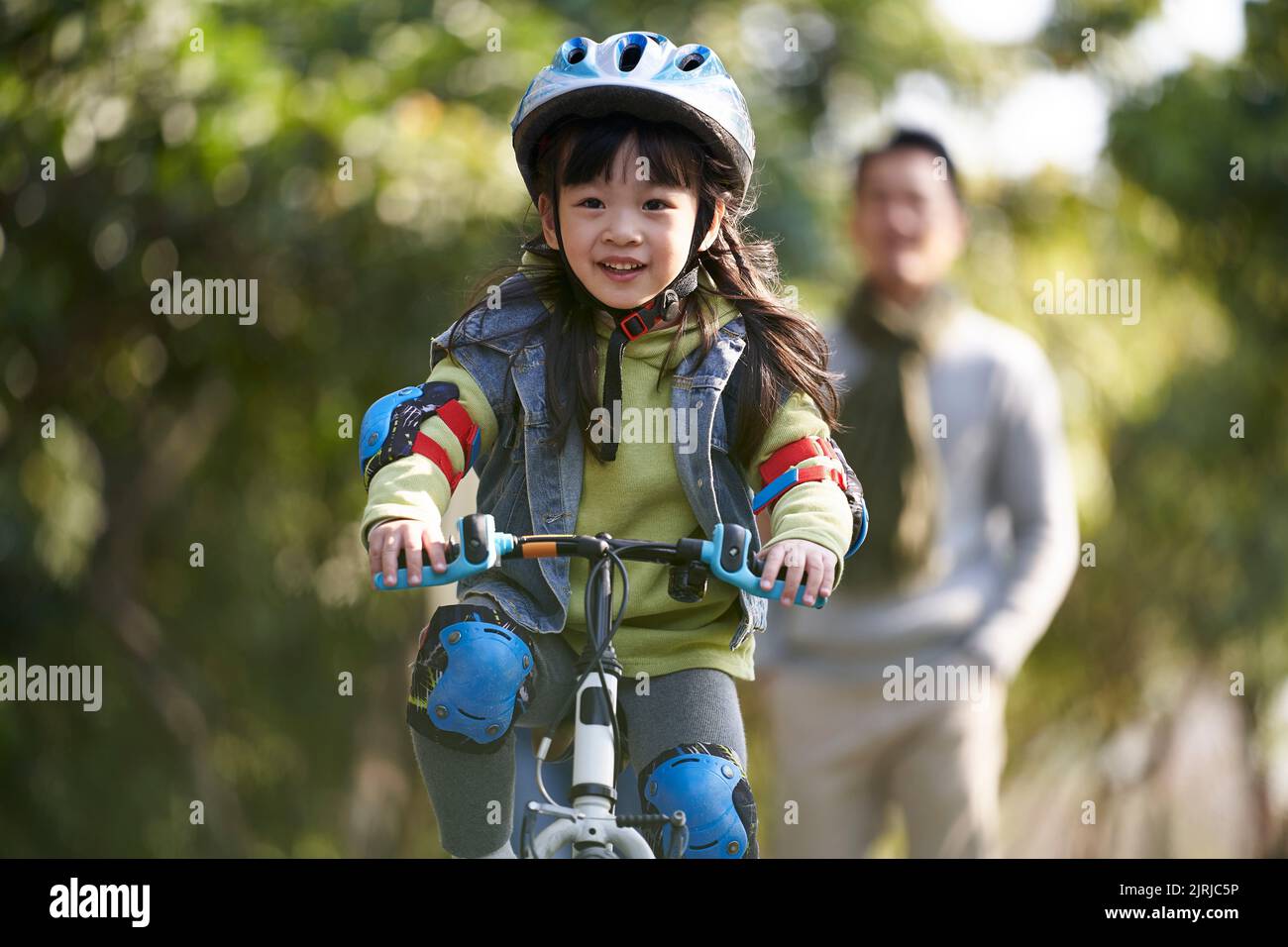little asian girl with helmet and protection gear riding bike in city park with father watching from behind Stock Photo