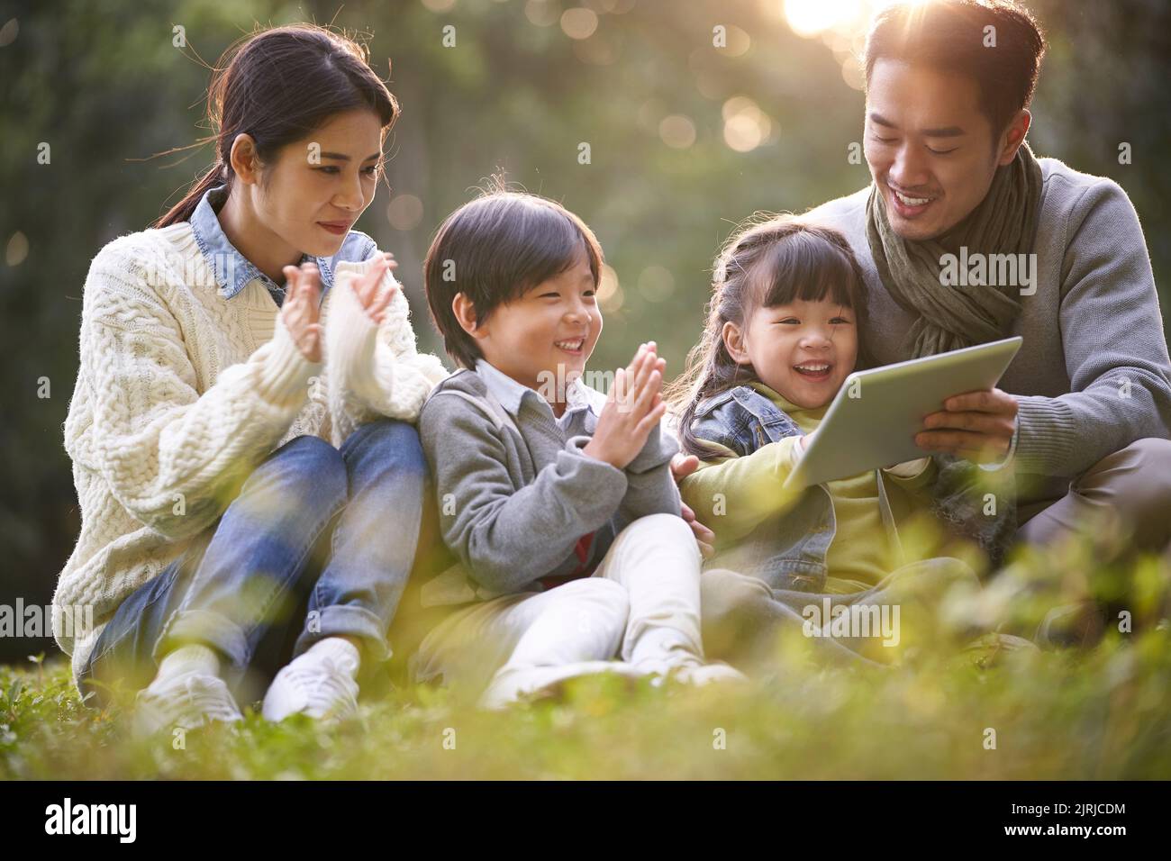 young asian family with two children sitting on grass outdoors in park watching contents in tablet computer Stock Photo