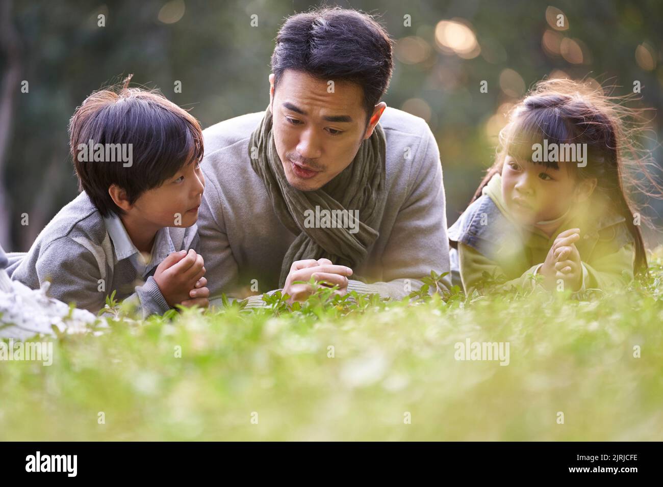 asian father lying on front on grass telling story to two children happy and smiling Stock Photo