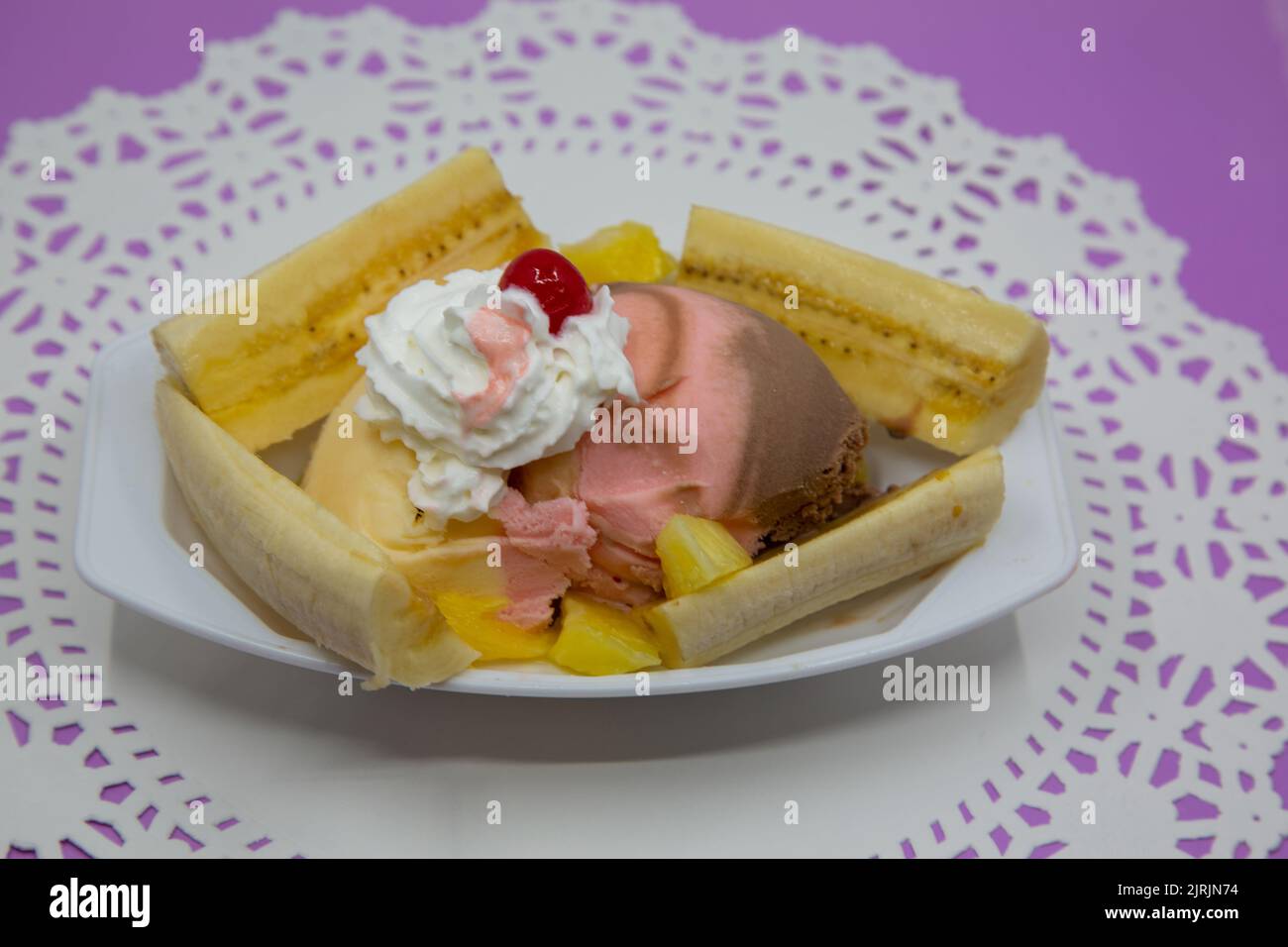 Banana split ice cream sundae on white doily and purple table top. Neapolitan ice cream with whipped creams, bananas, maraschino cherry and pineapple Stock Photo