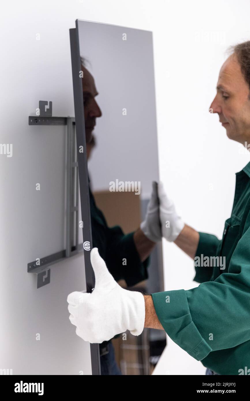 Tauberbischofsheim, Germany. 18th Aug, 2022. An employee of the manufacturer of infrared heaters Vitramo attaches an infrared heater for wall mounting to the wall. In an infrared heater, a frontal cover (glass or sheet metal) is heated from the back by a heating layer that converts electrical energy into heat. Credit: Daniel Karmann/dpa/Alamy Live News Stock Photo