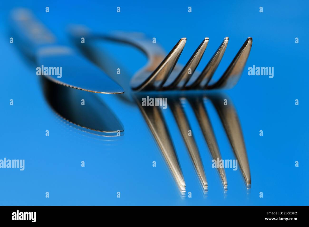 A knife and fork side-by-side resting on a mirror reflecting the undersides in a warmish blue mood light setting with only the ends in focus Stock Photo