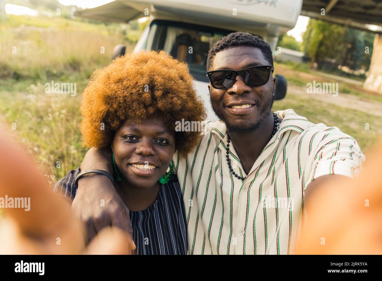 Smiling ethnic dark-skinned straight couple having a summer road trip in a camping van, hugging and taking a selfie in front of their vehicle. High quality photo Stock Photo