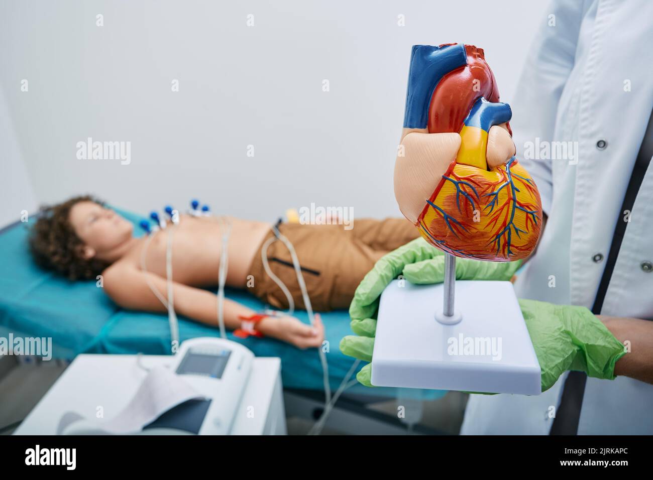 Anatomical model of human heart in doctor's hands over background of male child lying on bed during ECG procedure. Electrocardiography for children Stock Photo
