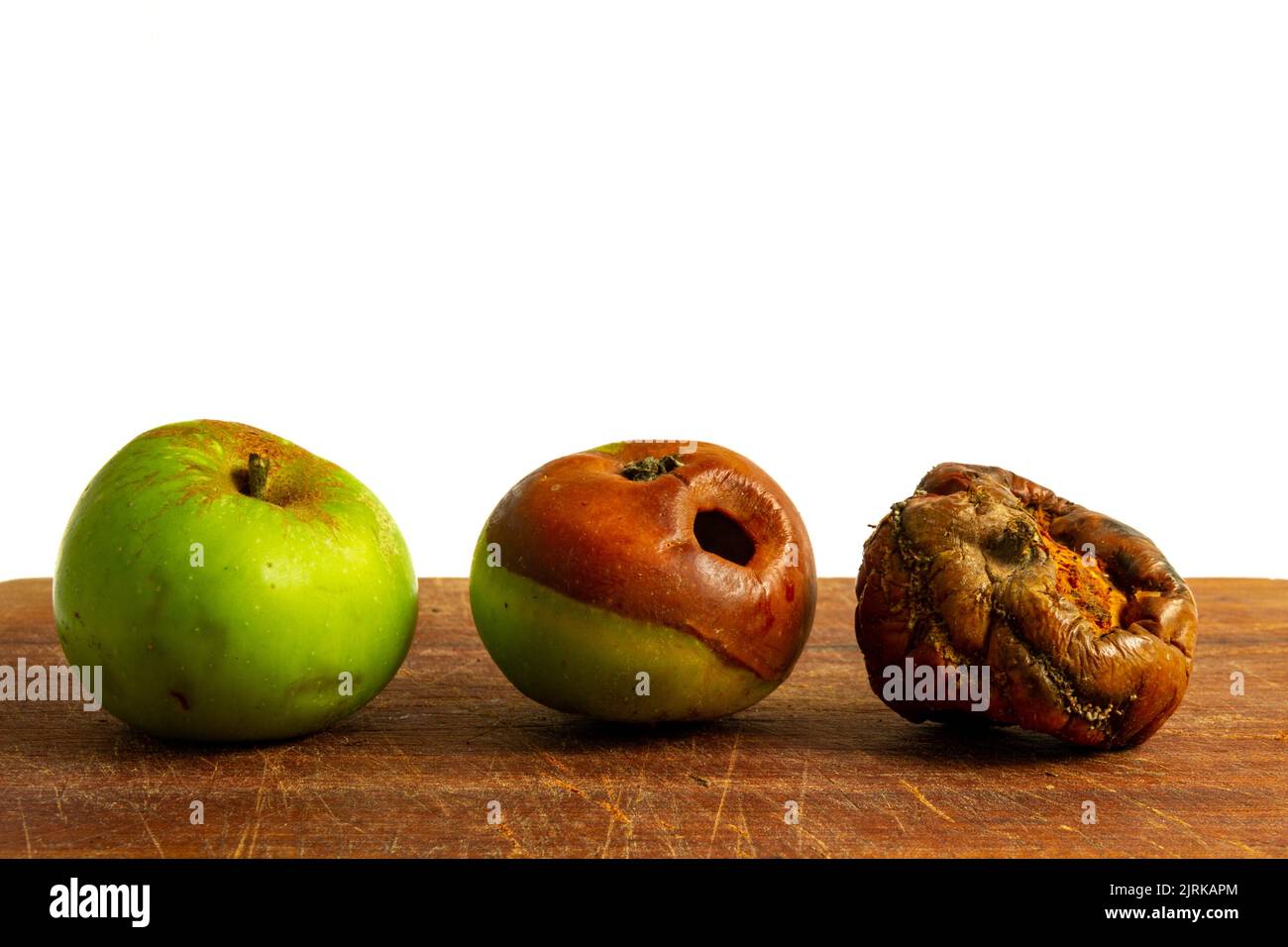 Three apples lined up on a wooden board against a white background, each of them in an increasing state of decay Stock Photo