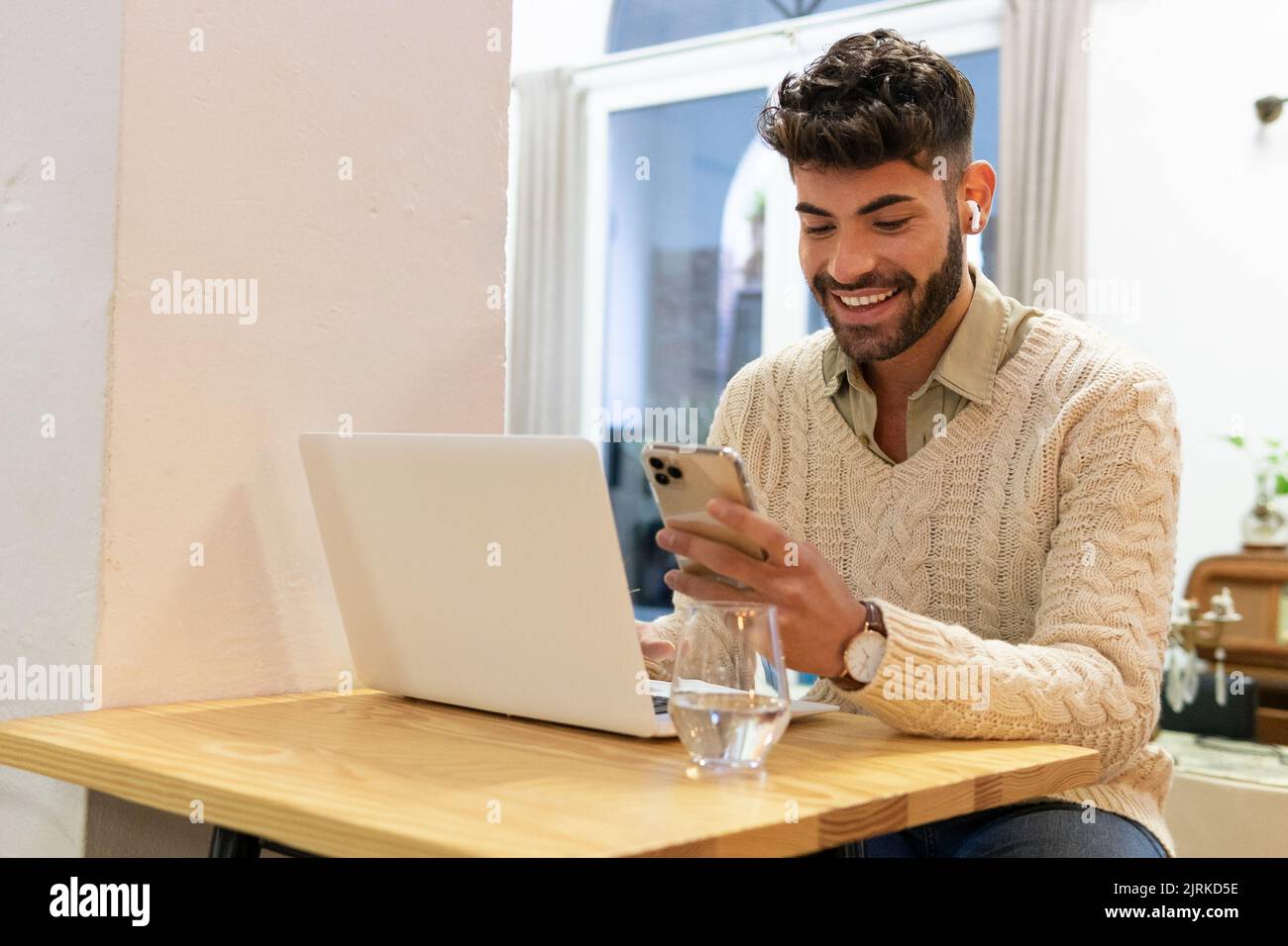 Smiling young Hispanic male with beard and dark hair sitting at home and browsing mobile phone Stock Photo