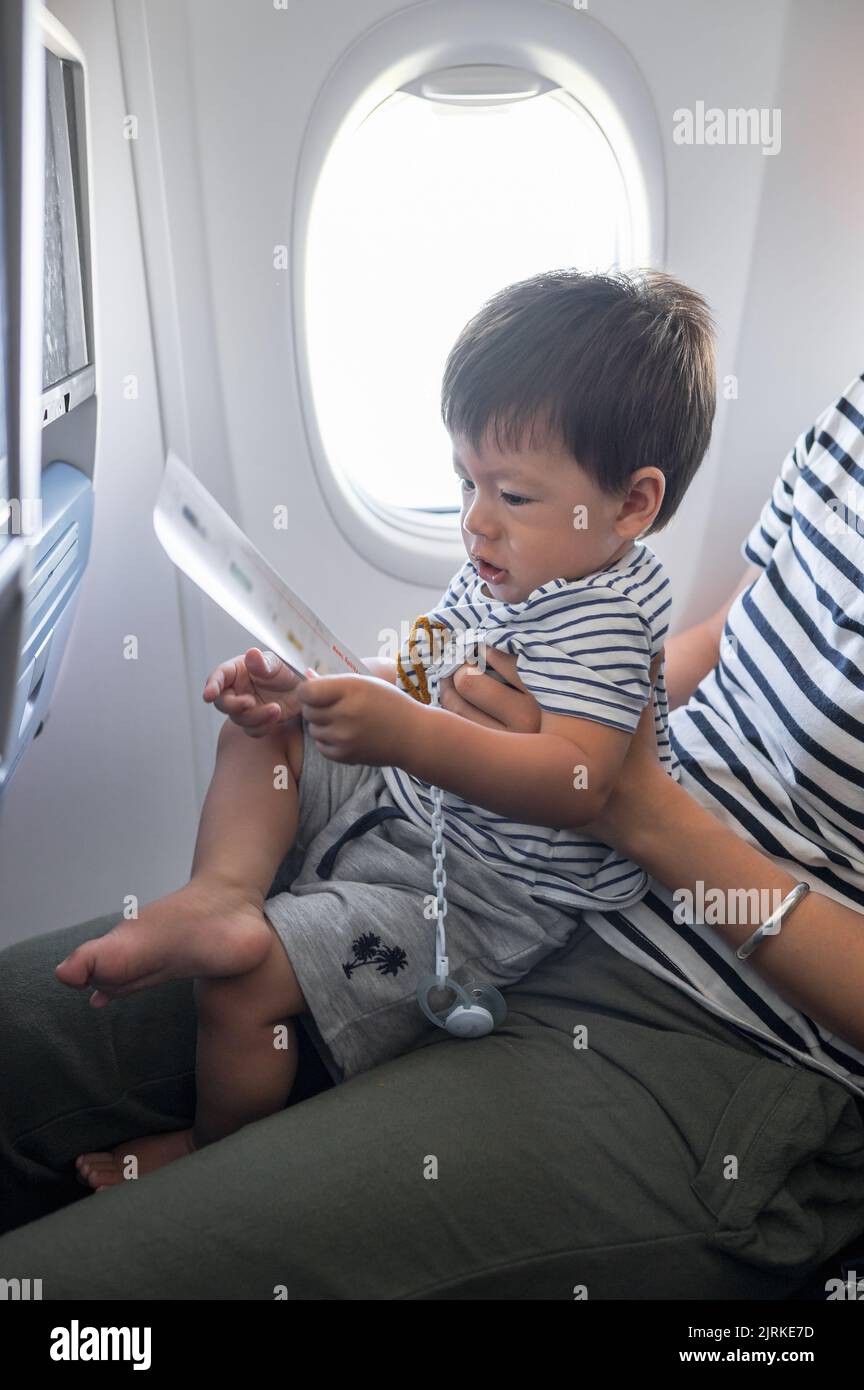 Infant traveling in airplane sitting on its mother lap. One year old baby boy flying in airplane and trying to entertain himself Stock Photo