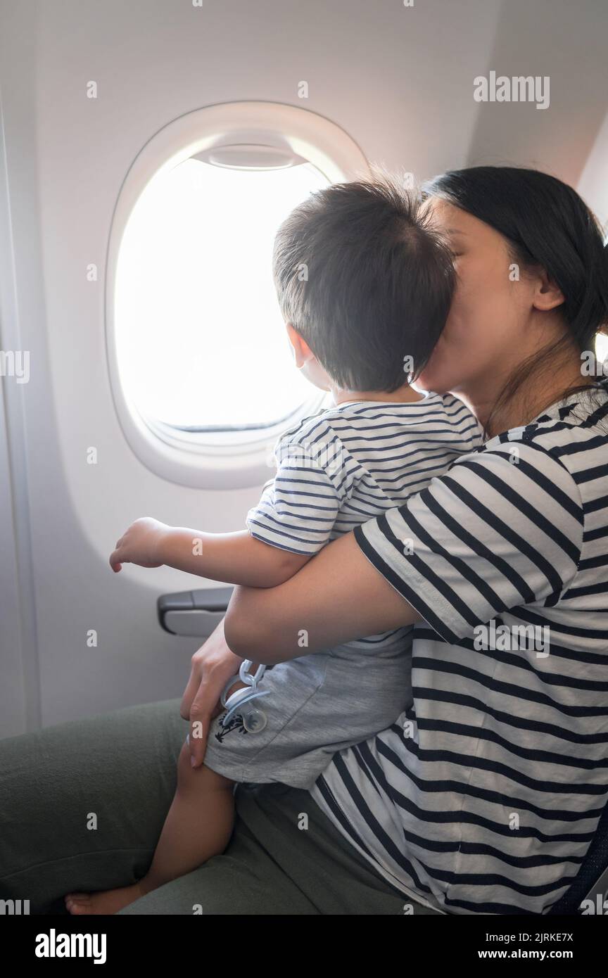 Infant traveling in airplane sitting on its mother lap both looking out of an airplane window. One year old baby boy flying in airplane and trying to Stock Photo
