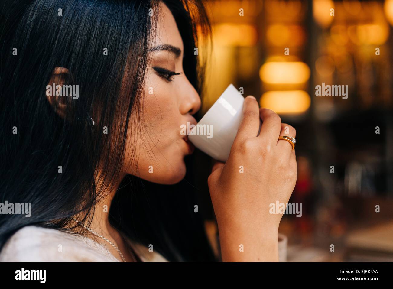 Side view of crop ethnic female with makeup drinking hot beverage from cup in coffee house Stock Photo