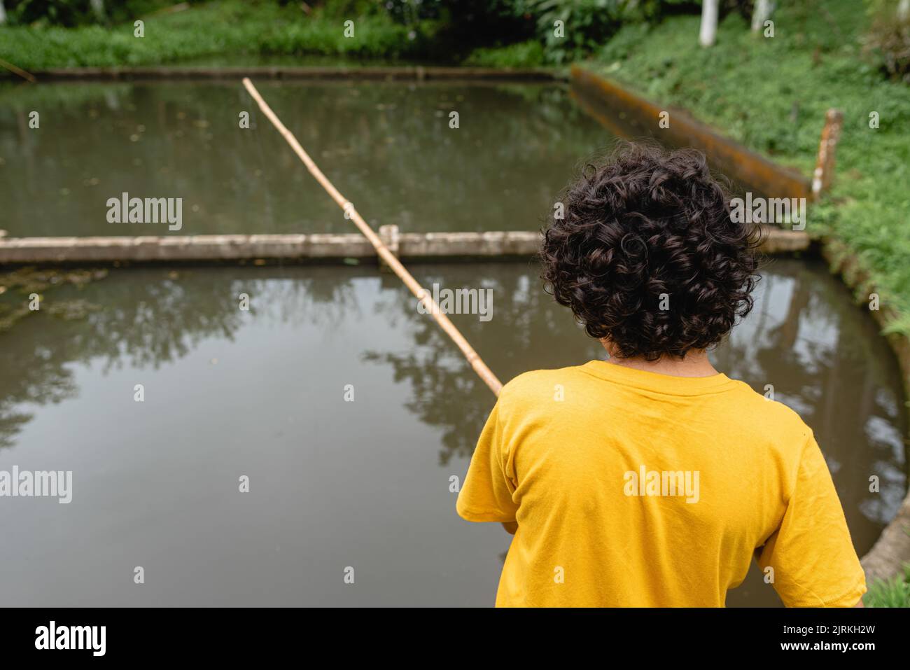 Back view of unrecognizable boy in casual clothes standing on shore near pond and fishing in green park in daytime Stock Photo