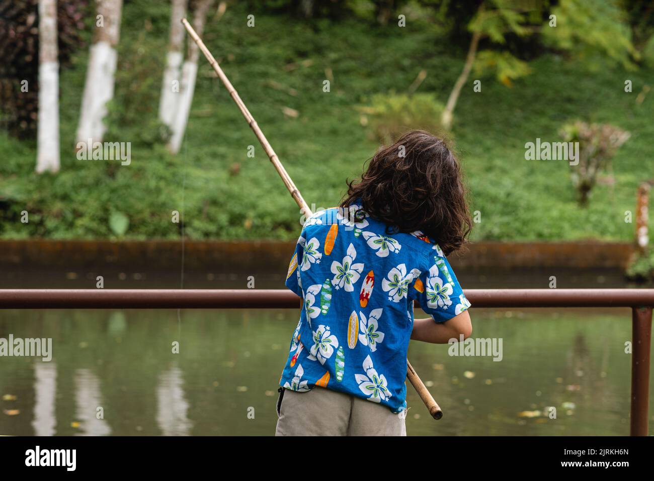 Back view of unrecognizable boy in casual clothes standing on shore near pond and fishing in green park in daytime Stock Photo