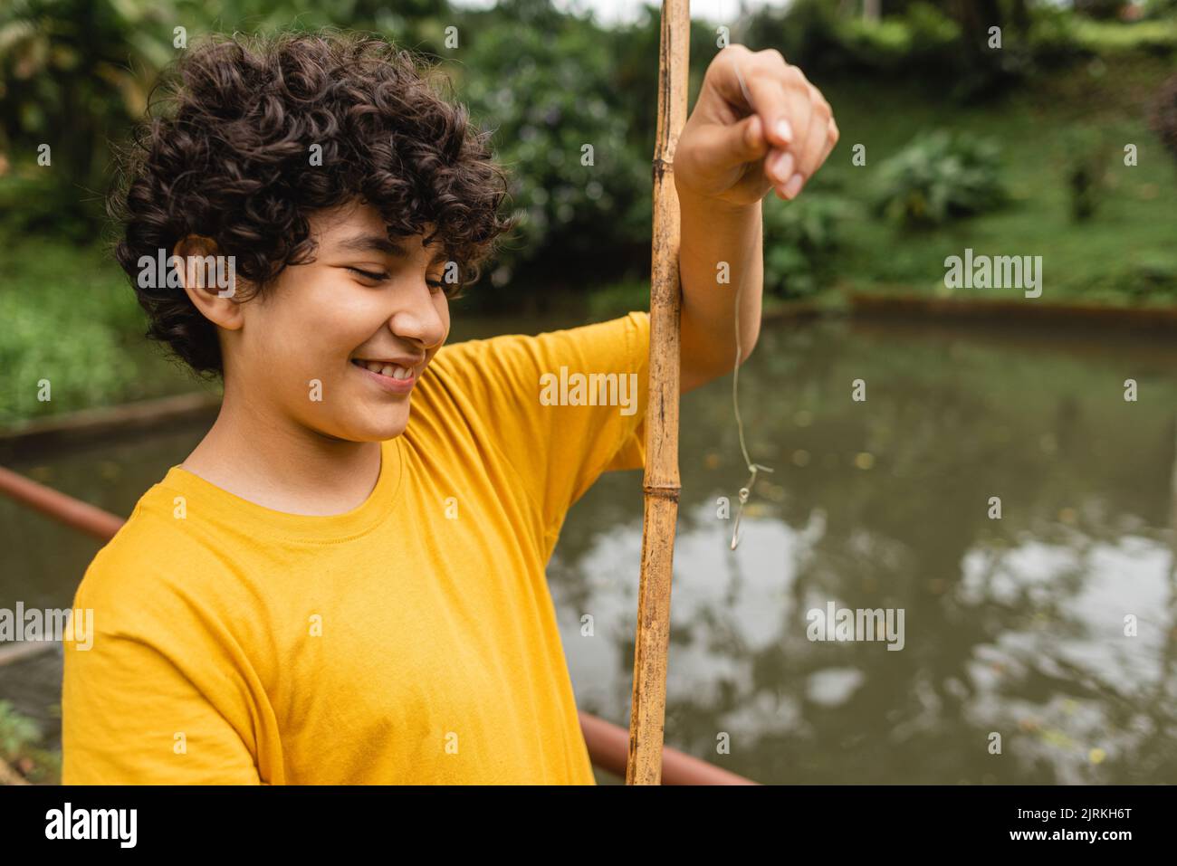 Side view of calm boy in casual clothes standing on shore near pond and fishing in green park in daytime Stock Photo
