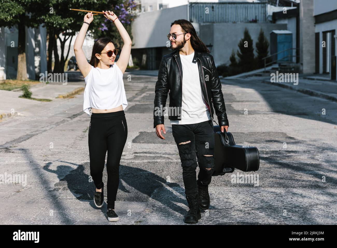 Smiling Latin American woman with drumsticks strolling on walkway against guitarist while talking and looking at each other in town Stock Photo