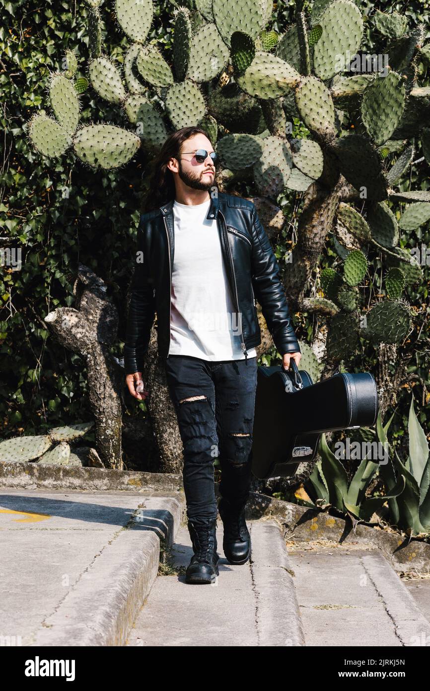 Trendy Latin American male guitarist in leather jacket and sunglasses carrying case with guitar while looking away on staircase against cactus leaves Stock Photo