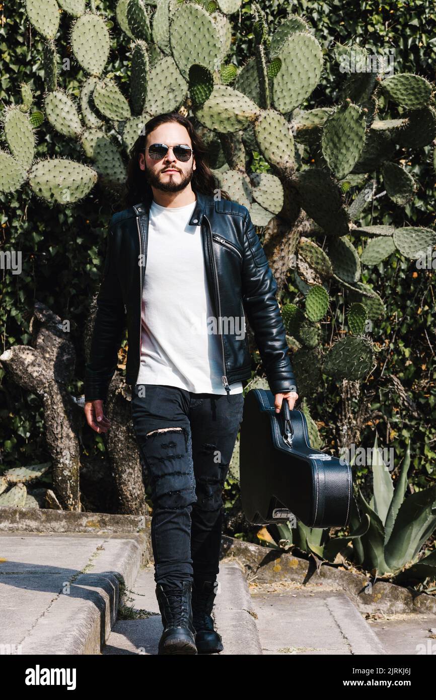 Trendy Latin American male guitarist in leather jacket and sunglasses carrying case with guitar while looking away on staircase against cactus leaves Stock Photo