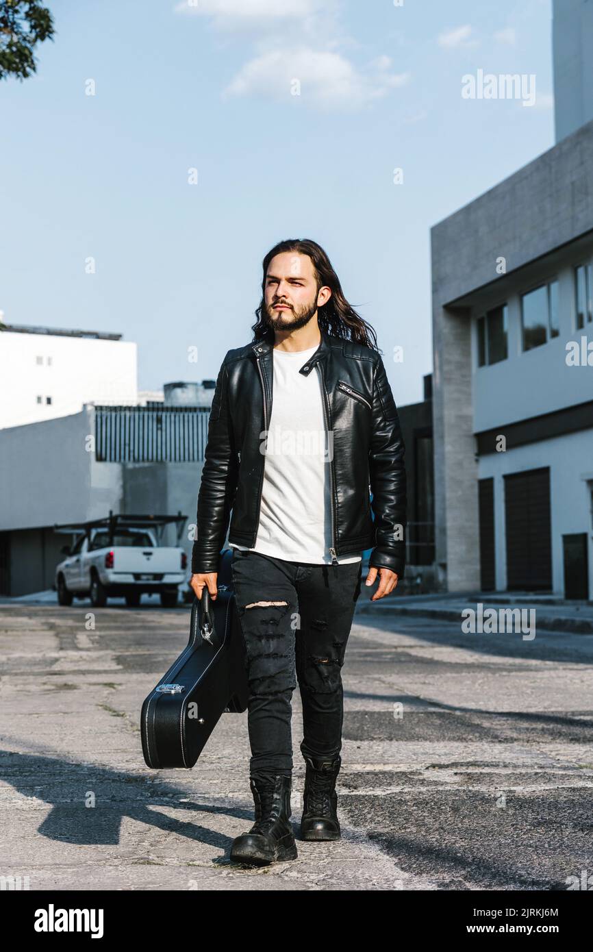 Trendy Latin American male guitarist in leather jacket carrying case with guitar while looking away on the street against buildings Stock Photo