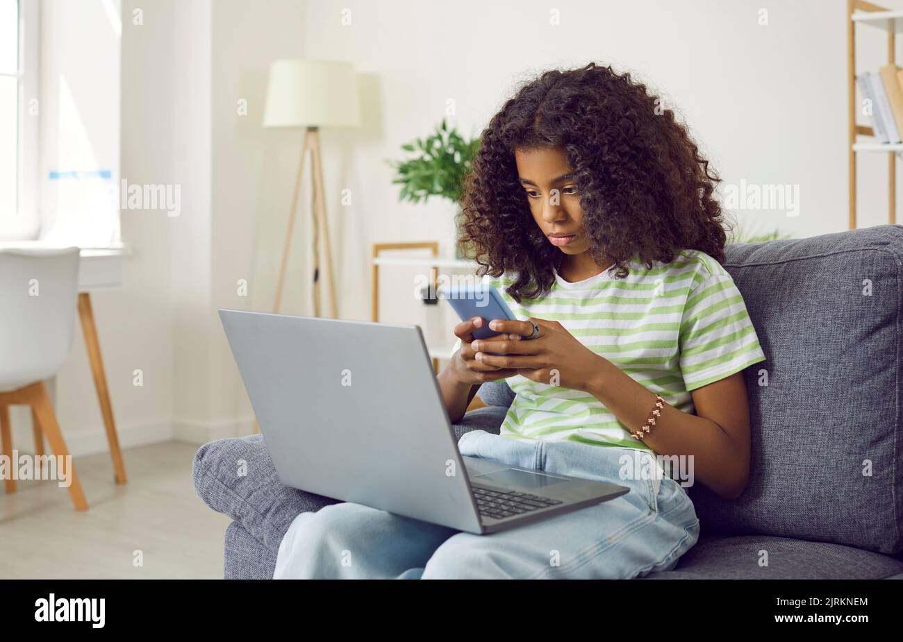 Young ethnic African American girl teenager uses laptop and phone at same time, sits on sofa at home Stock Photo