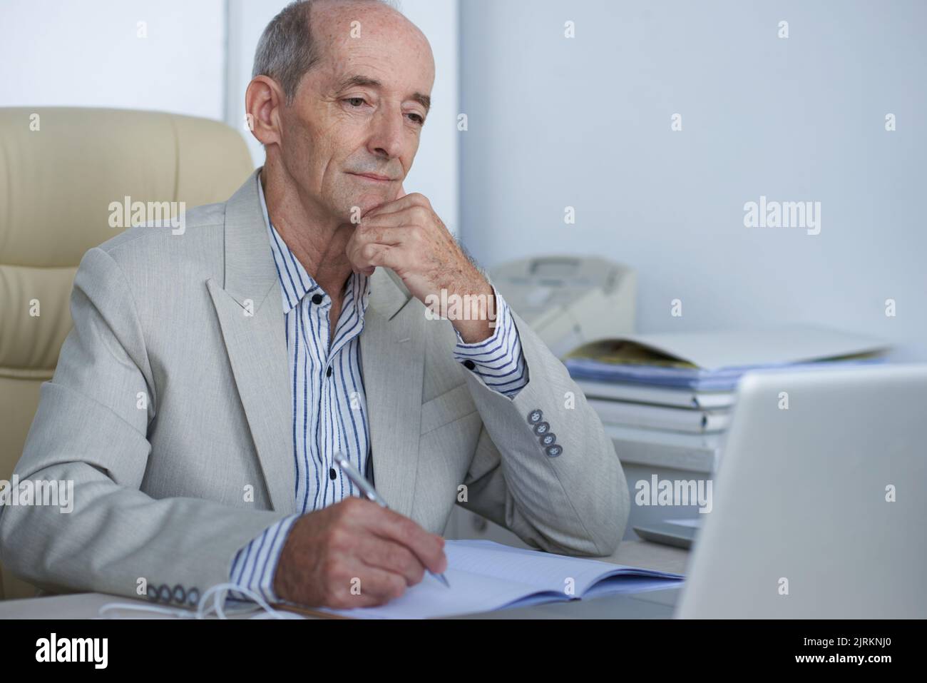 Pensive aged businesman reading something on laptop screen and taking notes Stock Photo