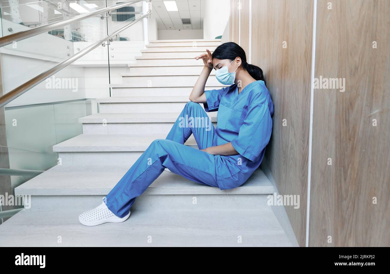 Tired female doctor after hard working day in medical clinic sitting on steps in hospital with eyes closed wearing medical mask. Pandemia coronavirus Stock Photo