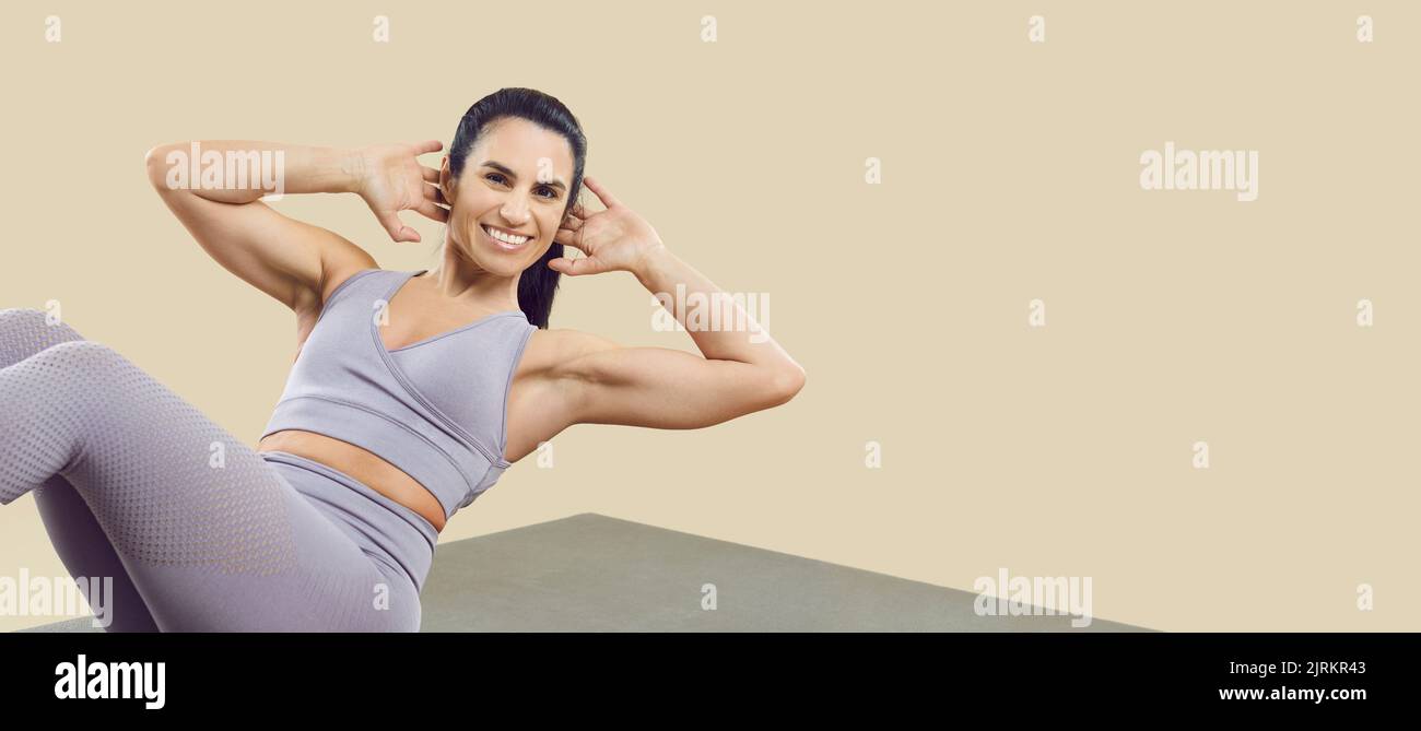 Smiling sporty woman doing abs exercises and crunches on sports mat isolated on beige background. Stock Photo