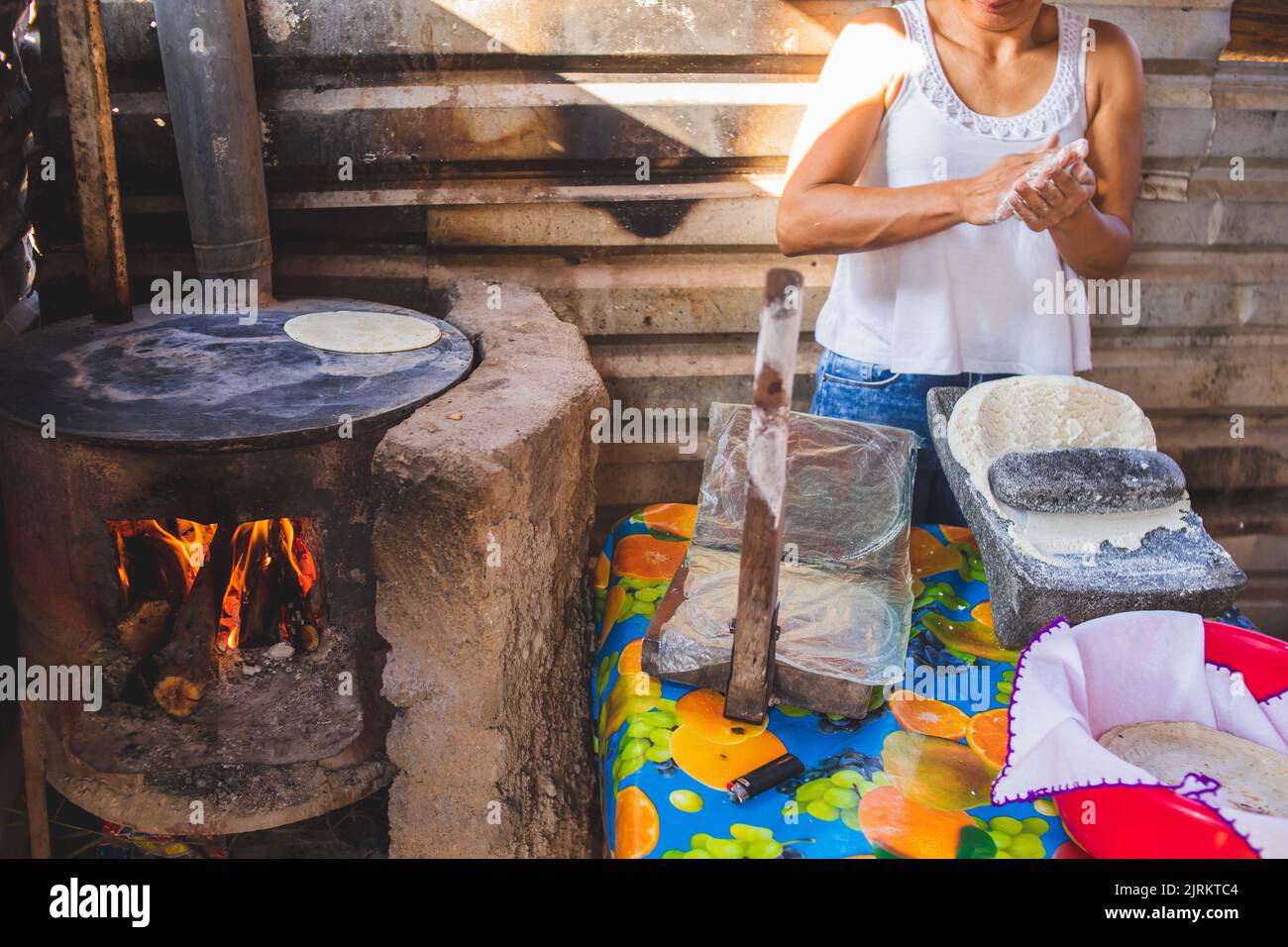 A Mexican woman torturing corn dough on a metate and a wood stove to make homemade tortillas Stock Photo