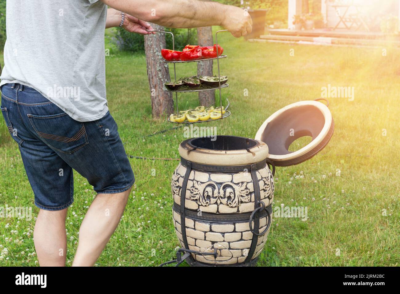 Cooking vegetables on the tandoor. Man holding a grill with vegetables, beautiful formal garden with dining place. Stock Photo