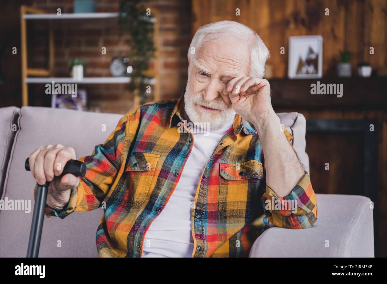 Photo of depressed stressed man pensioner wear checkered shirt holding walking cane crying sitting sofa indoors house room Stock Photo