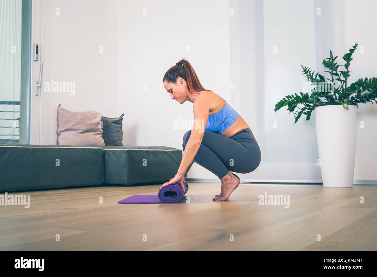 Young yoga woman rolling her violet mat after a yoga class on wooden floor near a window in her linving room Stock Photo