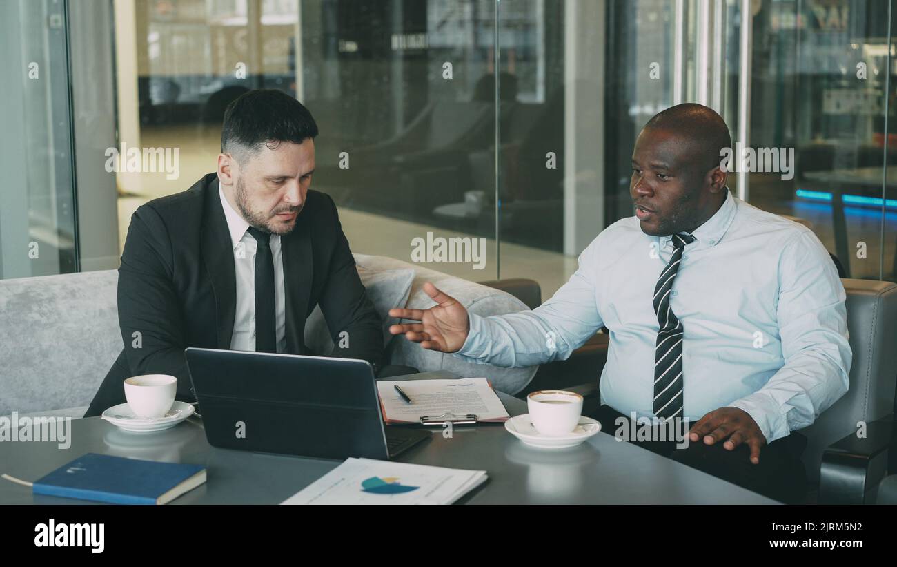 Angry businessman in shirt an tie criticizes severely his partner during meeting in modern cafe. Irritated african american boss gesticulates emotionally and agressively talking with colleague Stock Photo