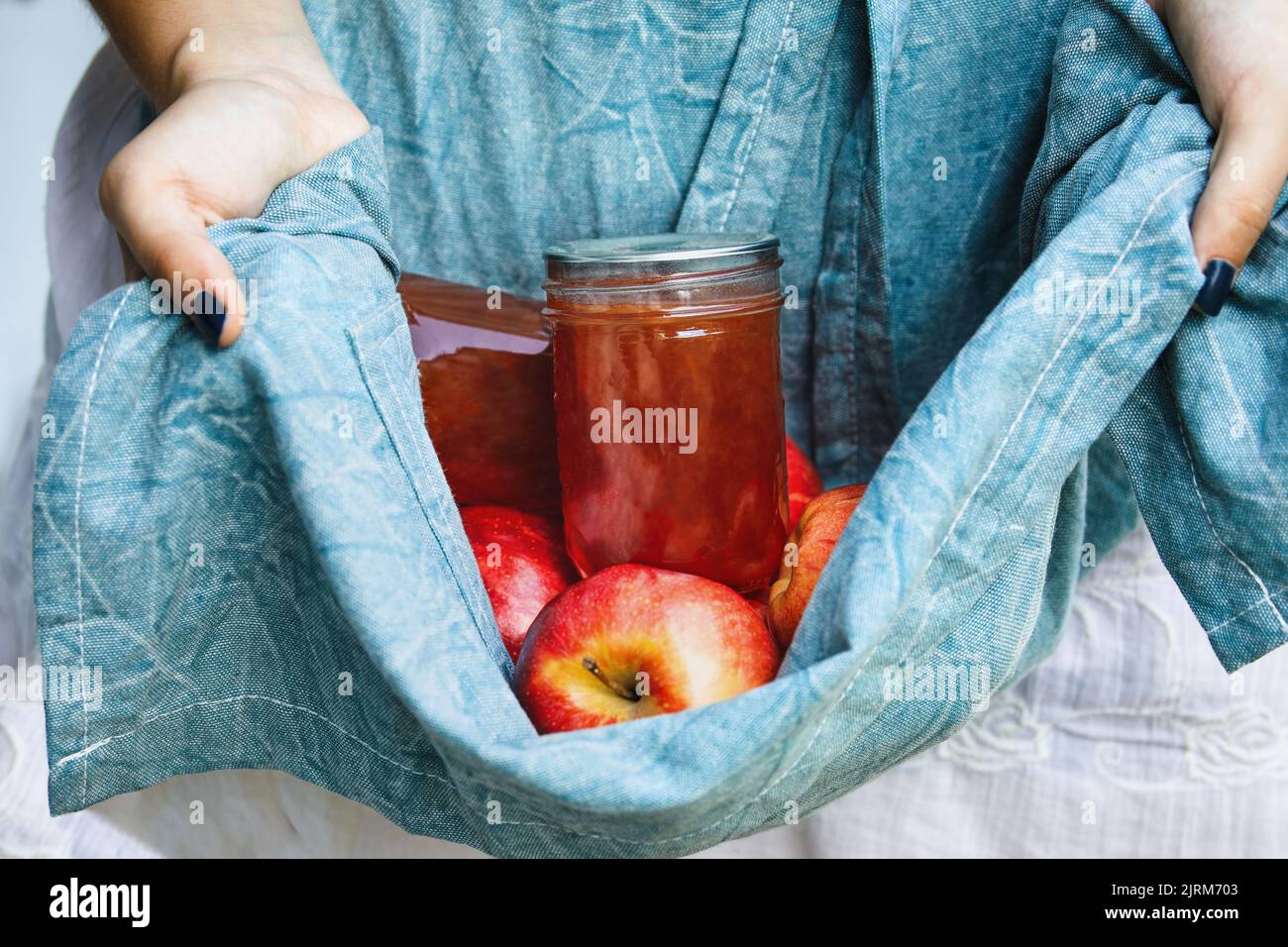 Woman's hands holding out apron filled with fresh gala apples and homemade apple jelly. Selective focus on mason jar with blurred background. Stock Photo