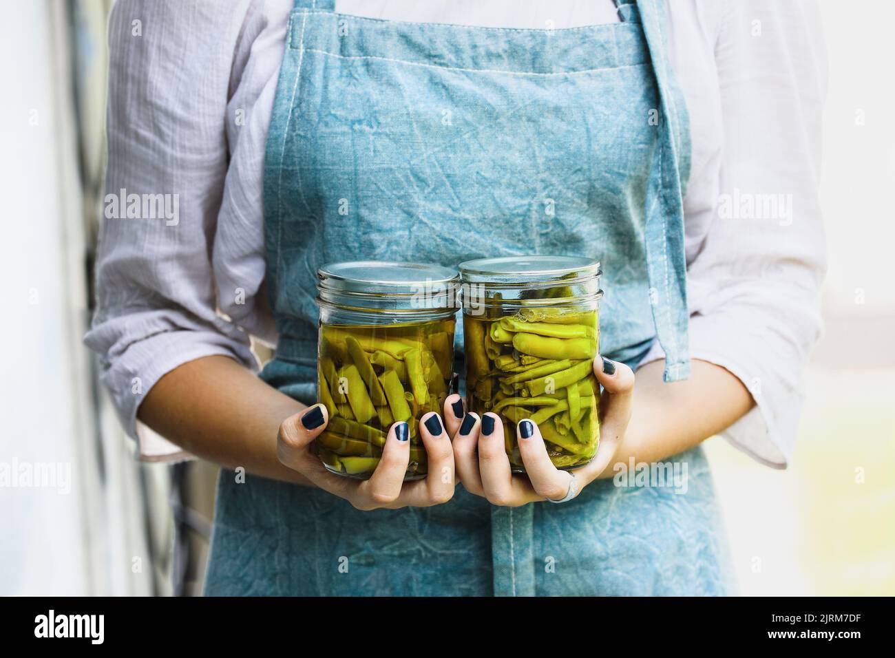 Young woman in a linen apron holding two mason jars filled with fresh home grown green beans. Selective focus blurred background. Stock Photo