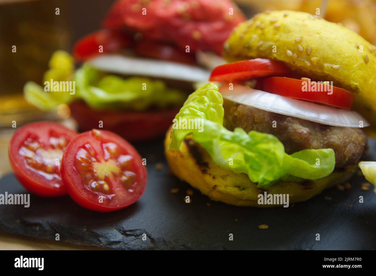Closeup of hamburgers with tomato, onion rings and lettuce and loaves of red and yellow colors, all on a black slate table Stock Photo