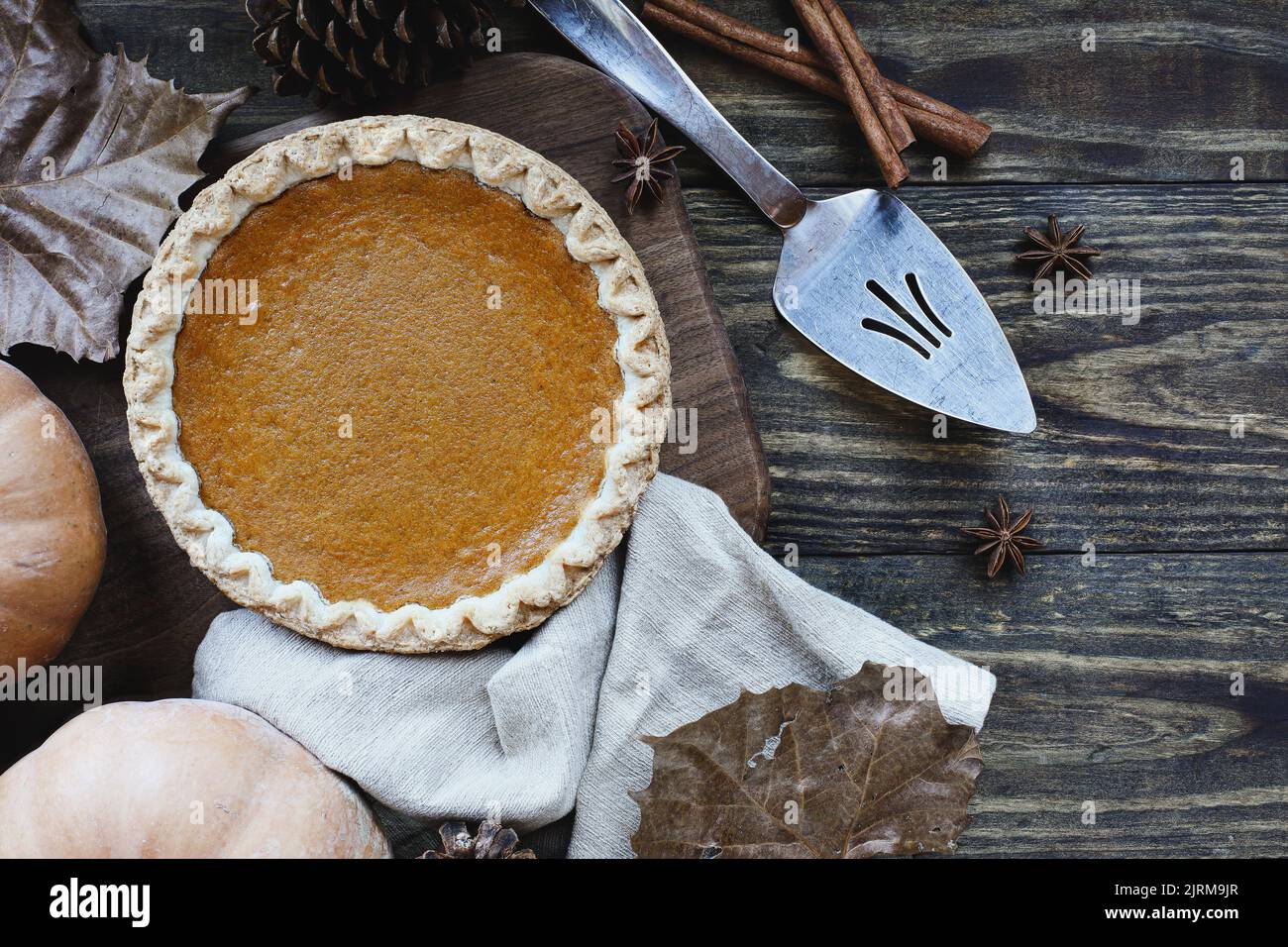 A whole fresh baked pumpkin pie over wood background with ripe pumpkins, cinnamon and star of anise. Top view or flatlay. Stock Photo