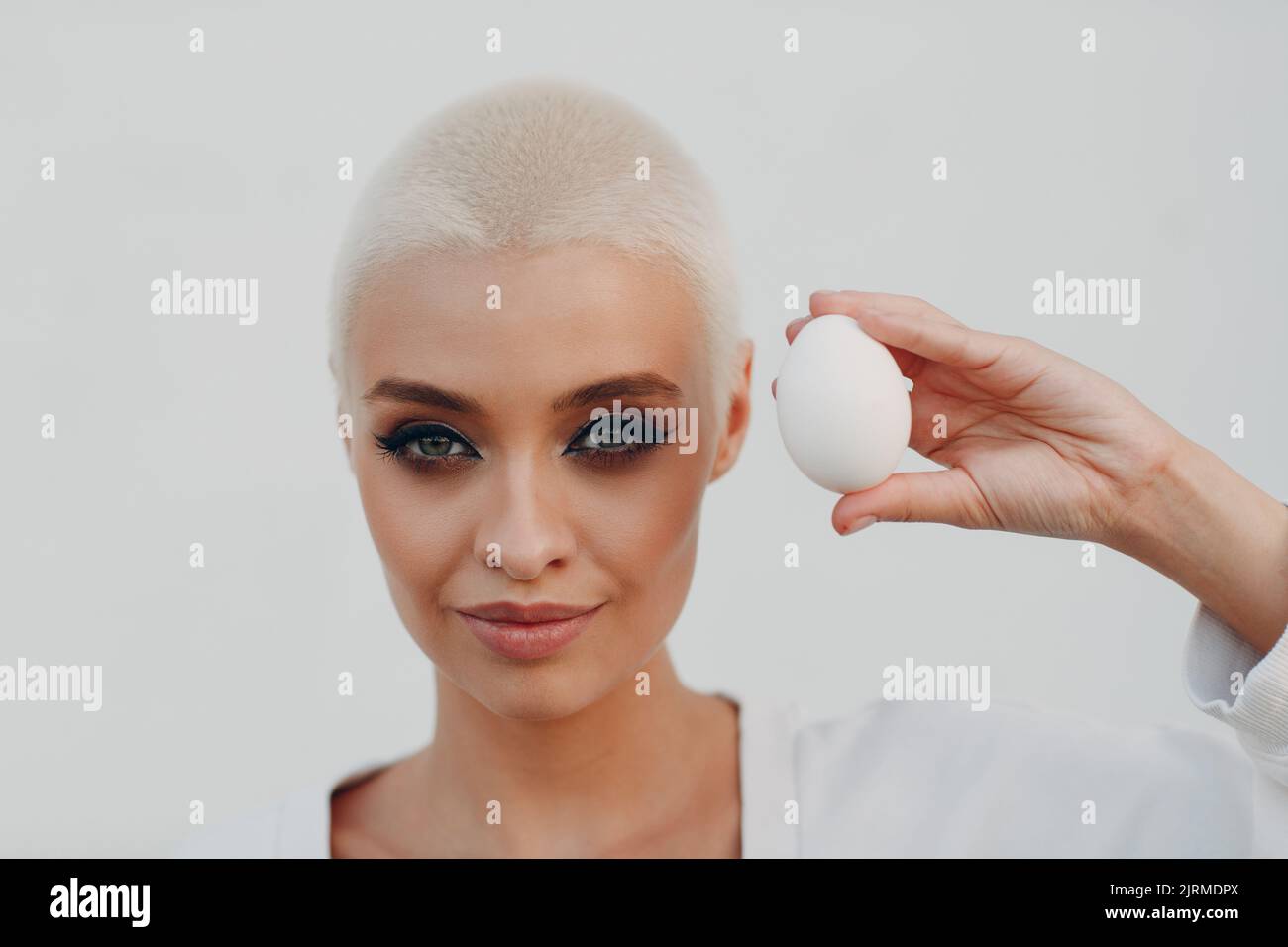 Portrait of young smiling millenial european short haired woman hold egg in hand. Beautiful happy blonde girl outdoor. Stock Photo