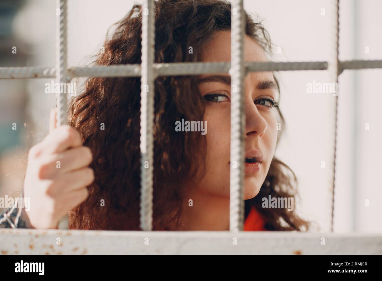 Young Brunette Curly Woman In Orange Suit Behind Jail Bars Female In Young Brunette Curly Woman In Orange Suit Behind Jail Bars Female In