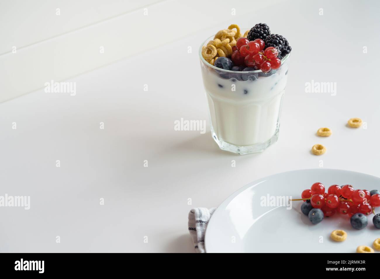 Yogurt, breakfast loops oat cereal,  and fresh summer berries fruits in glass on white table. Red currants, blueberries, blackberries on white plate, Stock Photo