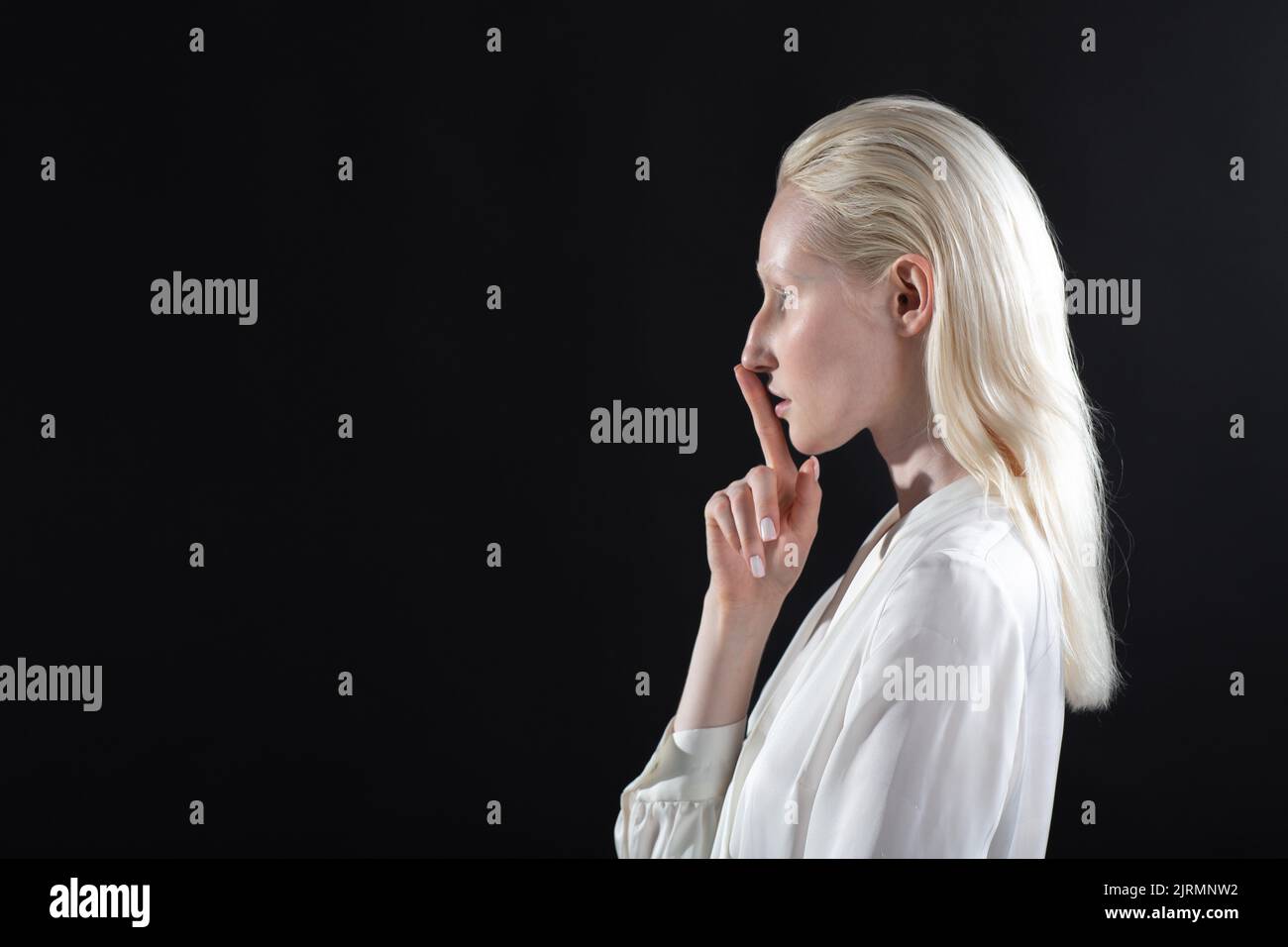 Closeup of beautiful young white caucasian albino blond woman with finger on her lips showing shhh silence gesture on black background. Stock Photo