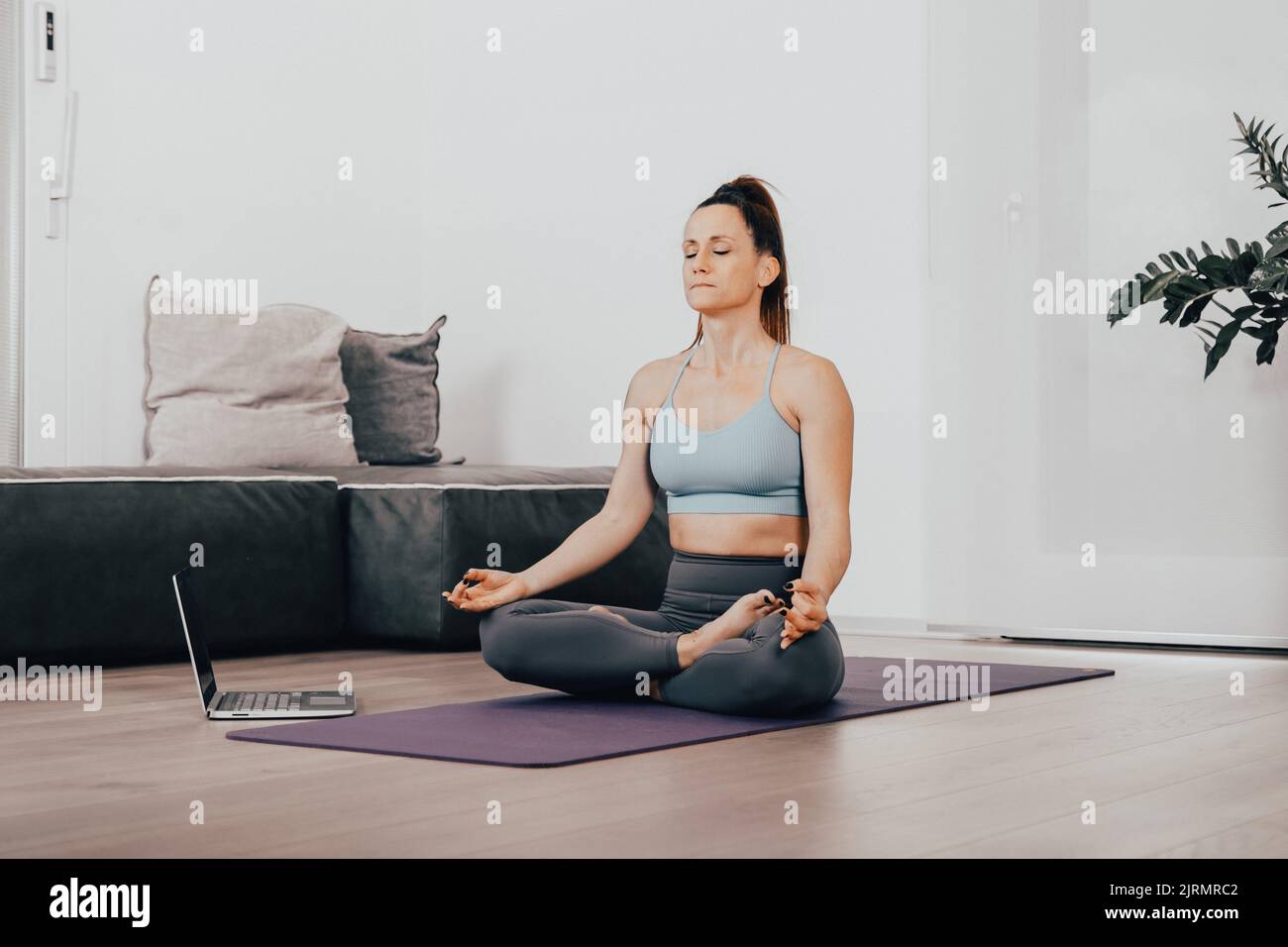 Calm woman  dressed in sportswear, meditates on the floor in her living room, listening to spiritual practices classes on laptop, poses in lotus pose, Stock Photo