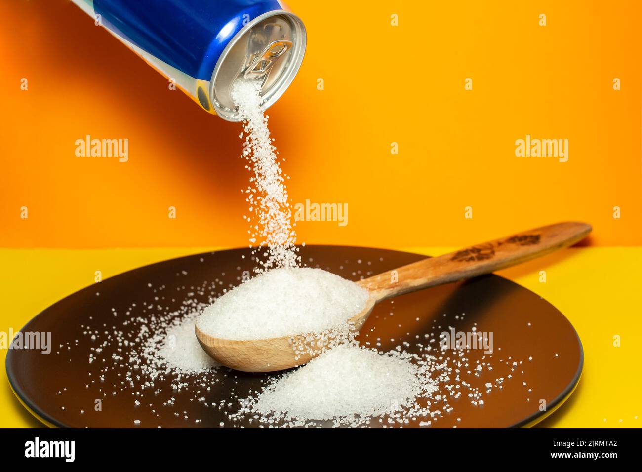 Pouring crystal sugar from a blue can to a wooden spoon on a black plate and orange background.  Motion blur on sugar texture. Sugar danger concept Stock Photo
