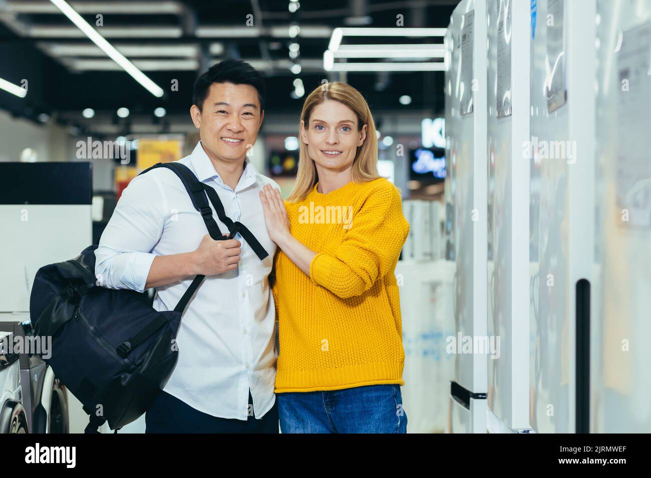 Portrait of a young married couple shopping in a home appliance supermarket, a diverse family of a man and a woman smiling and looking at the camera, choosing and buying home appliances Stock Photo