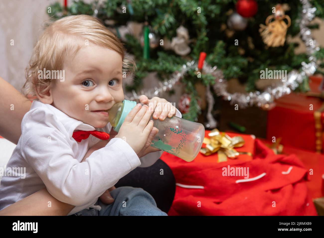 A little boy drinks from a bottle near a Christmas tree with gifts