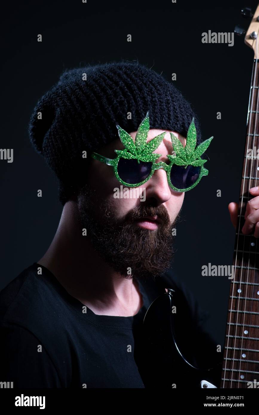 portrait of a bearded musician guy, wearing marijuana sunglasses, emotionally playing the electric guitar. on a dark background. Stock Photo