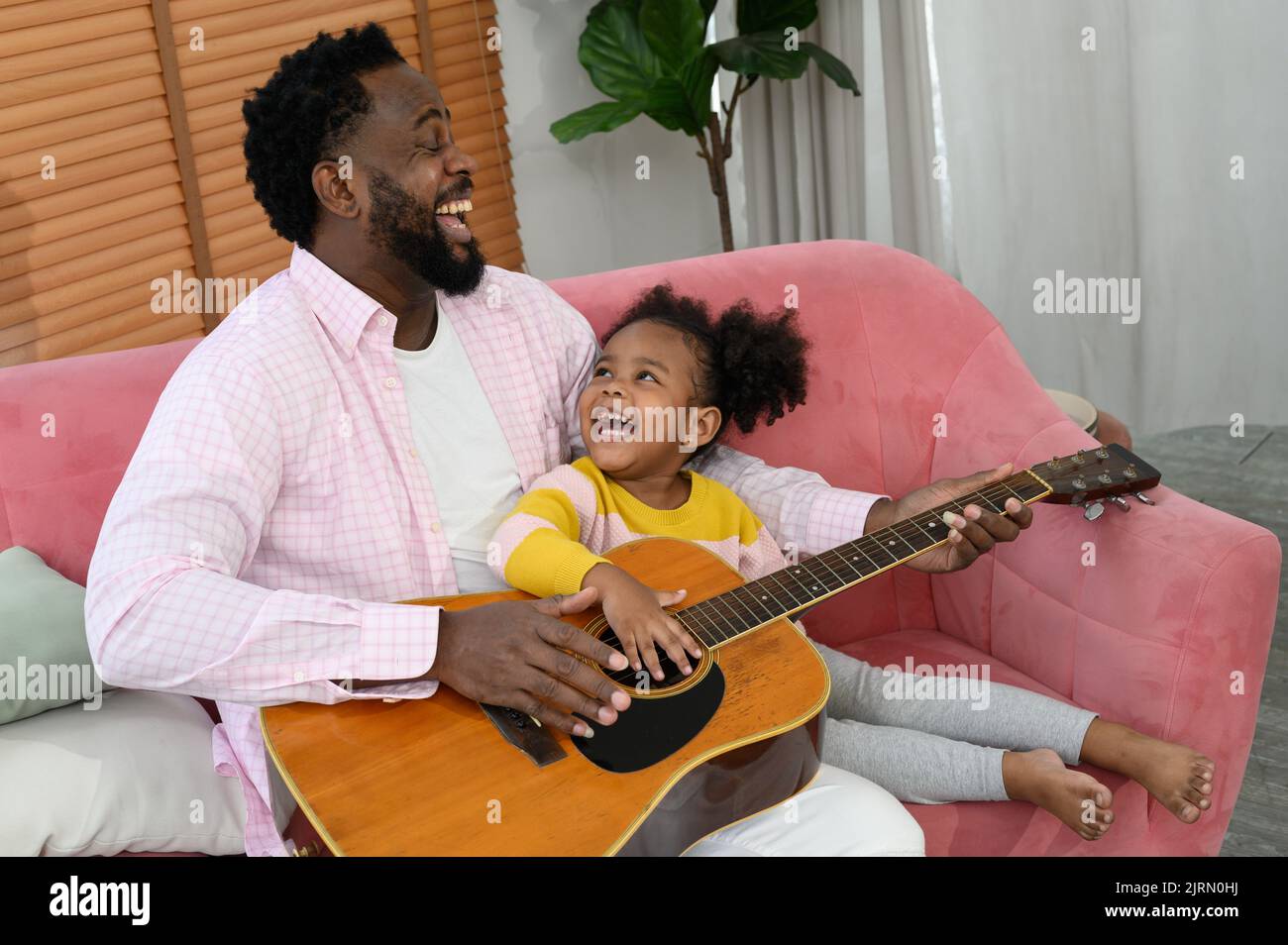 African girl and father are playing guitar and smiling while sitting on couch Stock Photo