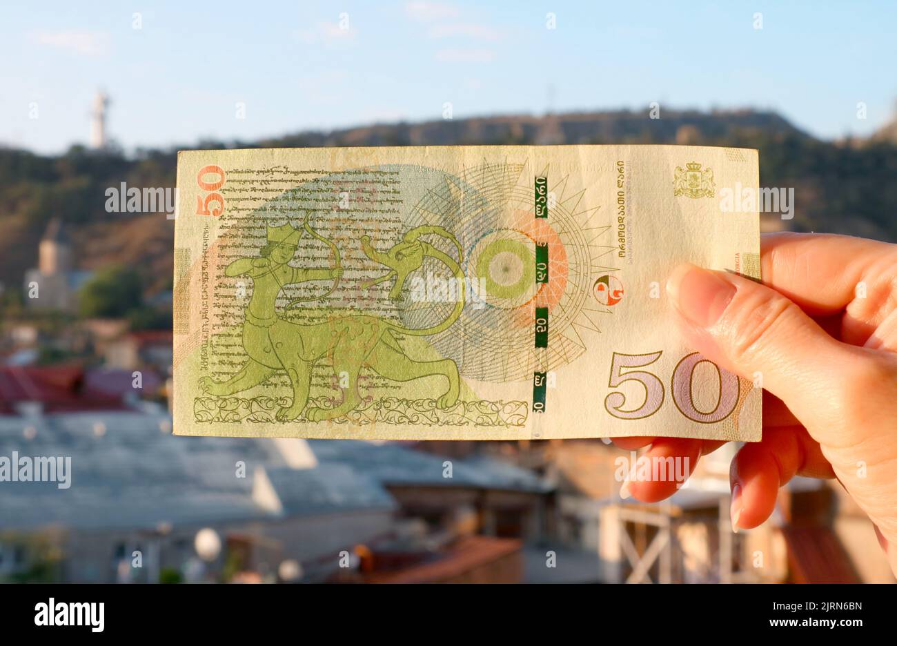 Hand Holding 50 Georgian Lari Banknotes in Reverse Side with Blurry Tbilisi City View in Backdrop Stock Photo