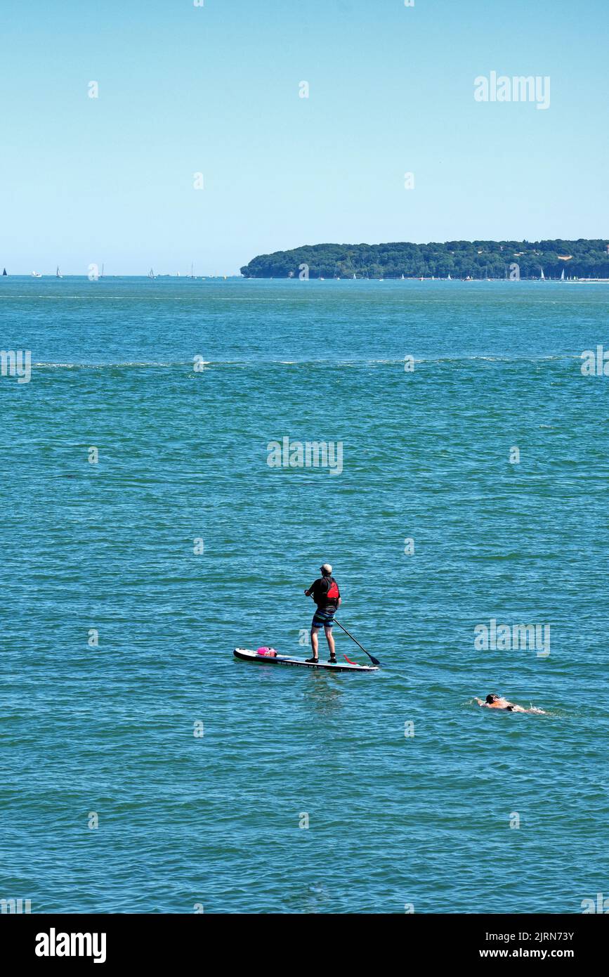 A man paddle boarding  with a swimmer next to him on the blue sea at Lepe on a summers day Hampshire England UK Stock Photo
