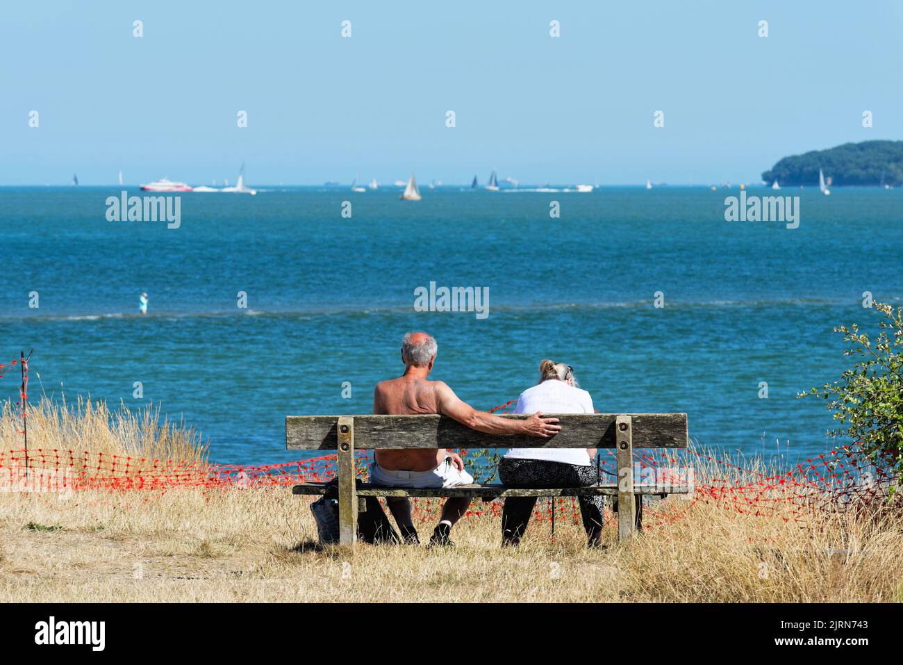 Rear view of an elderly couple sitting on a bench enjoying the view across the Solent on a summers day at Lepe country park, Hampshire England UK Stock Photo