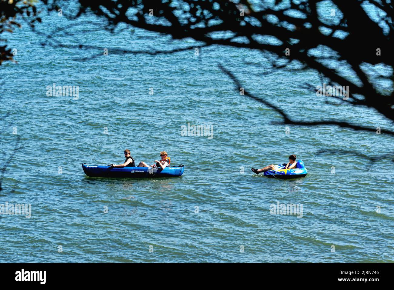 Two teenage boys in a canoe towing a third boy in an inflatable ring in the sea at Lepe beach on a hot summers day Hampshire England UK Stock Photo